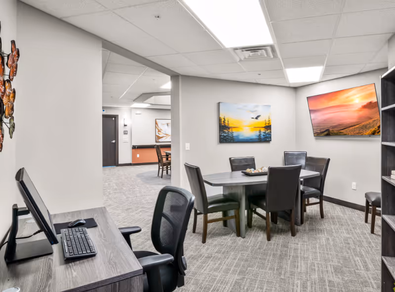 Interior common area with a round table and chairs, a computer workstation, wall-mounted TV and artwork on the walls.