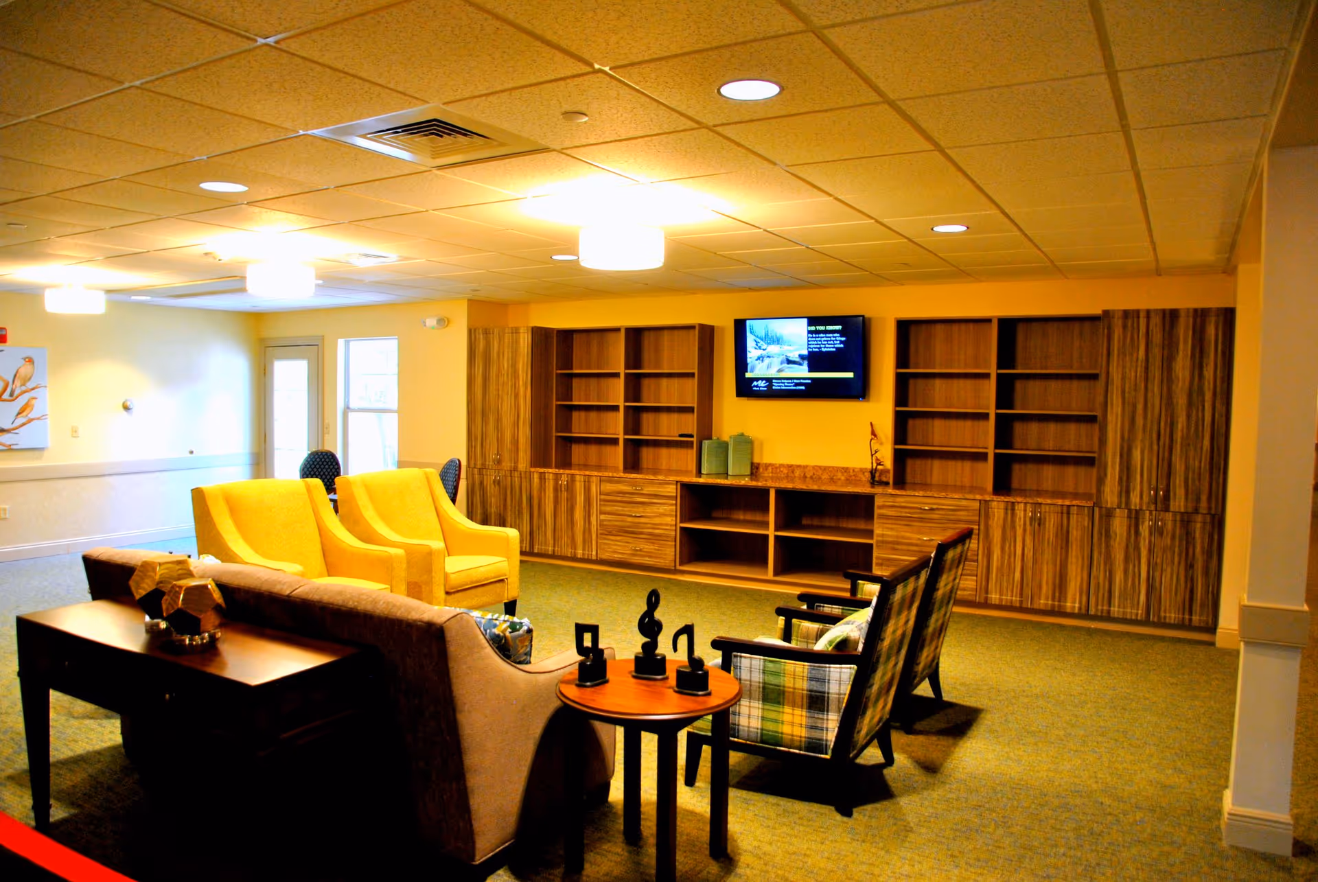A cozy living room area in an assisted living facility featuring a brown sofa, two yellow armchairs, and two plaid armchairs arranged around a small round wooden table with decorative musical note sculptures. The room has built-in wooden shelves and cabinets along the back wall, a wall-mounted TV, and soft overhead lighting.