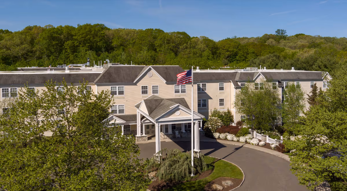 A senior living facility exterior with a central entrance, flag, and landscaped grounds.