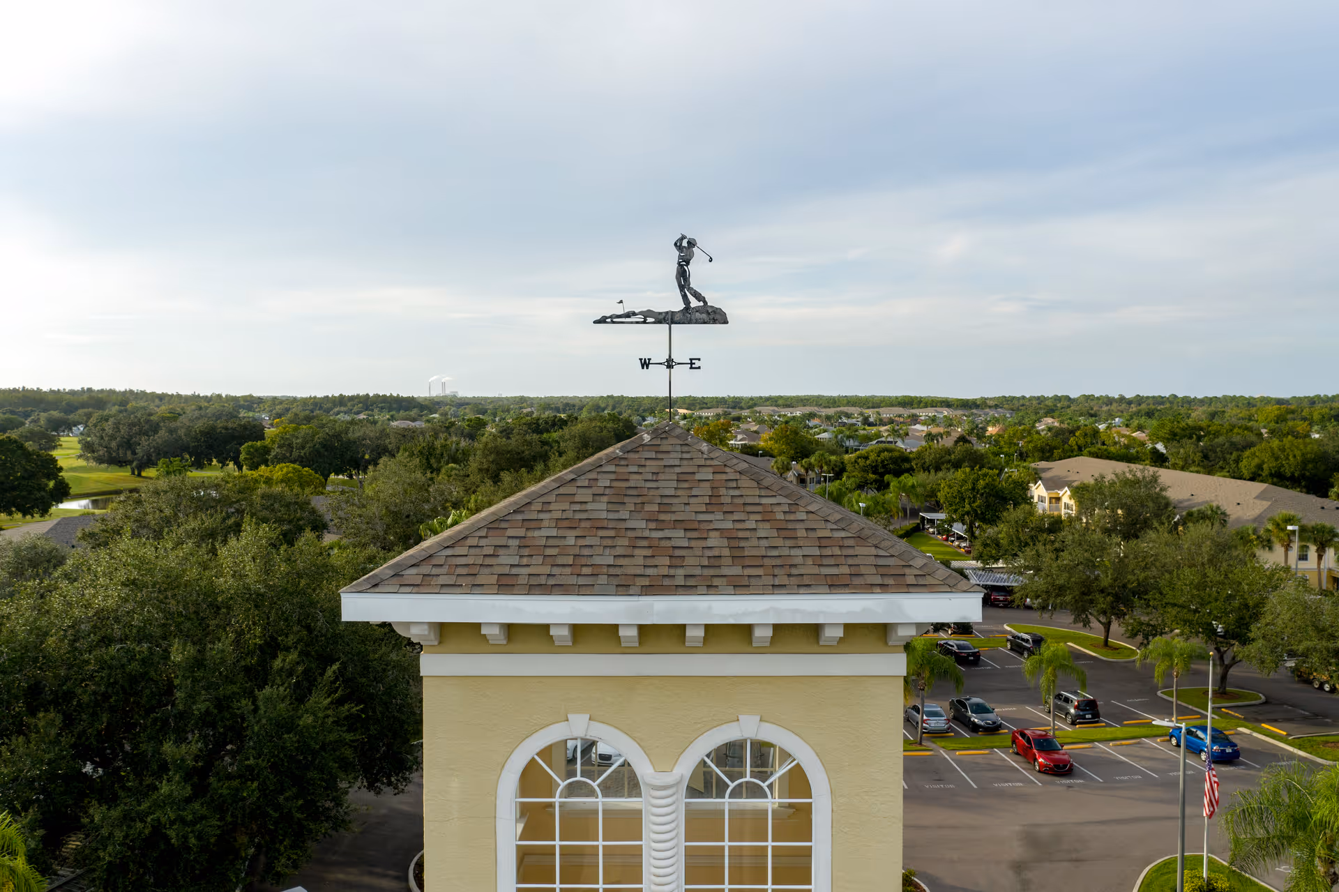 Aerial view of the roof of a building with a weather vane featuring a golfer figure on top. Surrounding the building are trees, parked cars, and other buildings under a partly cloudy sky.
