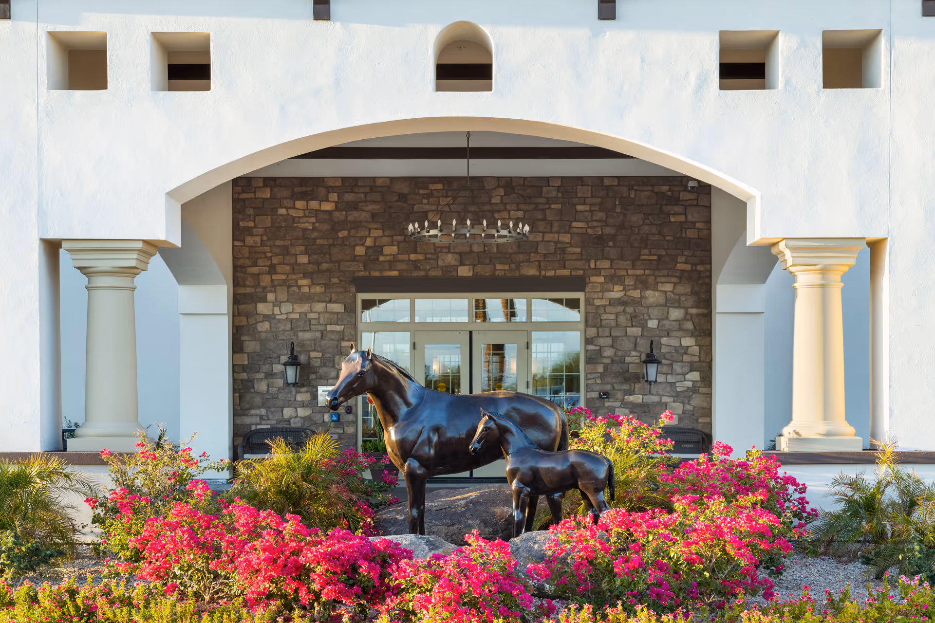 Bronze horse and foal statues in a flowerbed in front of a stucco building entrance with columns and an arched portico.