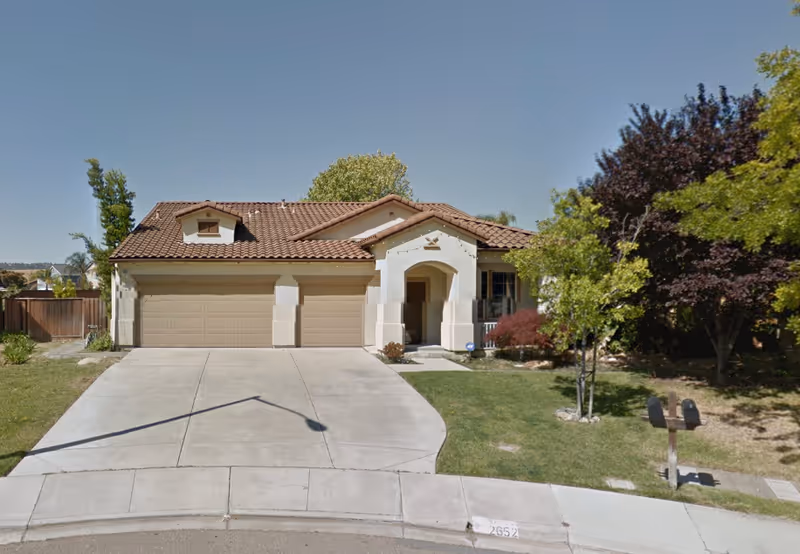 Front exterior of a single-story stucco house with a three-car garage, driveway, lawn, and trees.