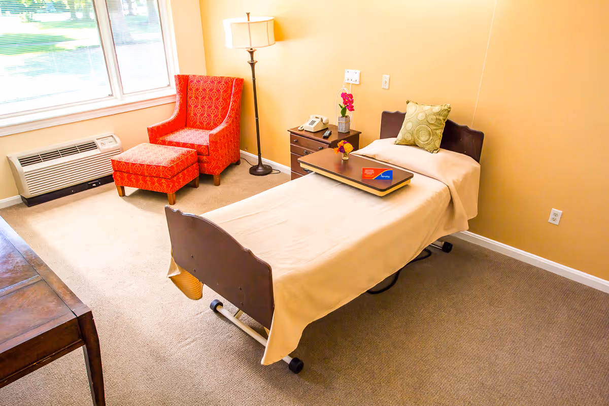 A bright and clean room with a single bed covered in a beige blanket and a green decorative pillow. Next to the bed is a small wooden nightstand with a telephone, a small vase with pink flowers, and a lamp. There is a red patterned armchair with a matching ottoman near a large window with blinds. The room has beige walls and carpeted floor.