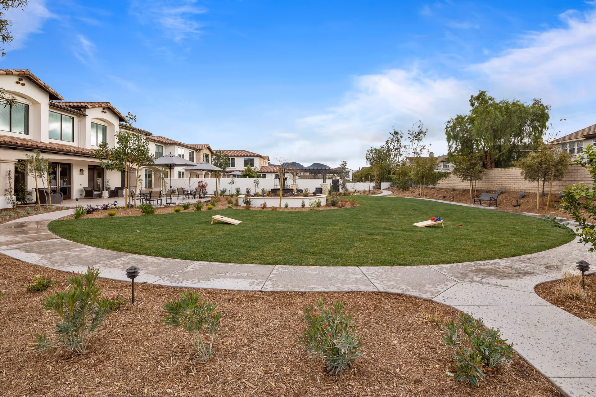 Outdoor garden area at Oakmont of Camarillo featuring a large circular grassy lawn with two cornhole boards, surrounded by a curved concrete walkway. There are patio tables with umbrellas and chairs along the building side, small trees and plants around the lawn, and a pergola structure in the background under a partly cloudy blue sky.