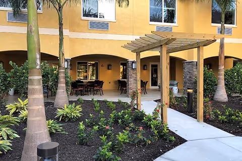 Outdoor patio area at Aravilla Sarasota with a wooden pergola over a concrete walkway, surrounded by landscaped garden beds with small plants and palm trees. The building exterior is yellow with arched openings leading to a covered seating area with tables and chairs.