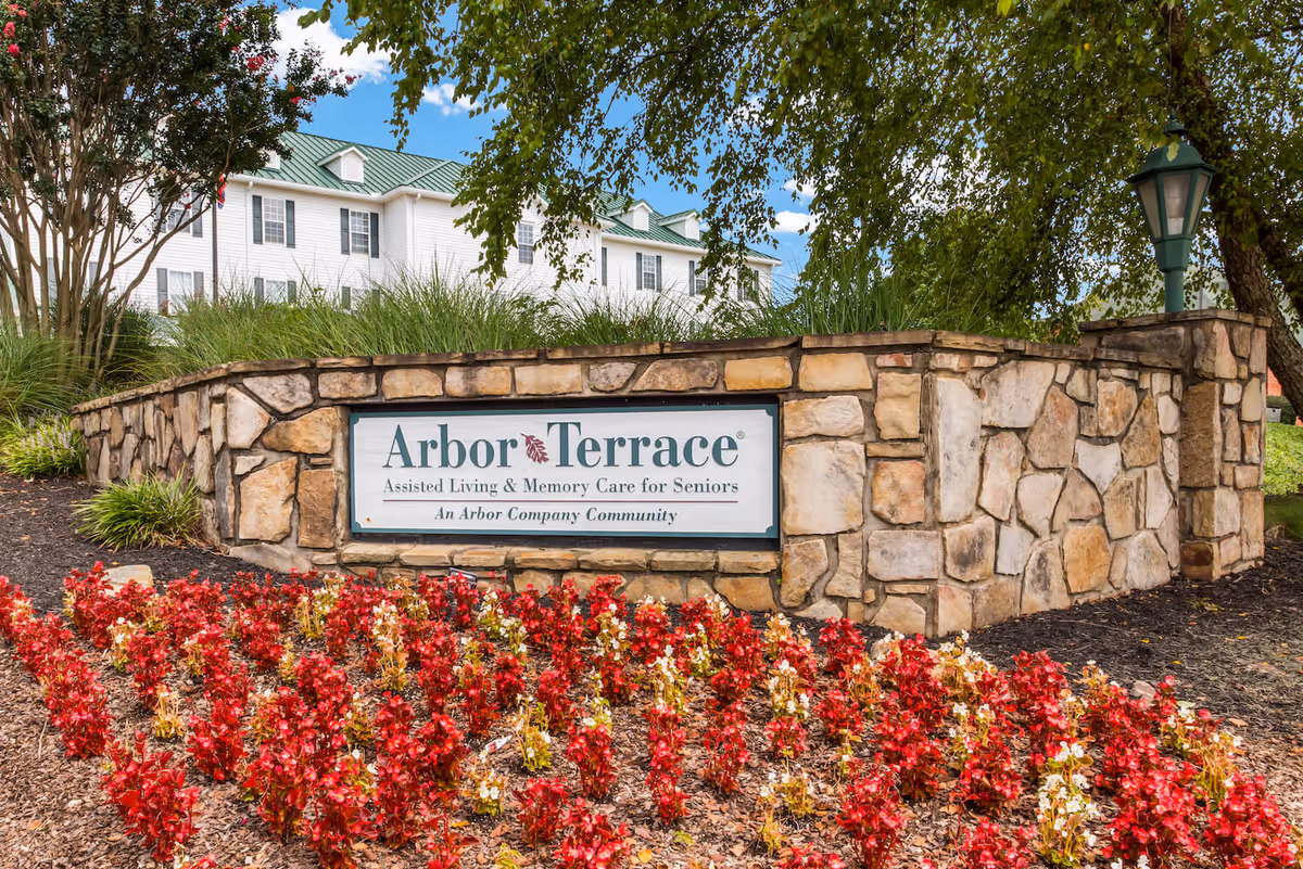 Stone sign for Arbor Terrace Assisted Living & Memory Care for Seniors surrounded by red and white flowers, with a white building and green roof in the background under a blue sky.