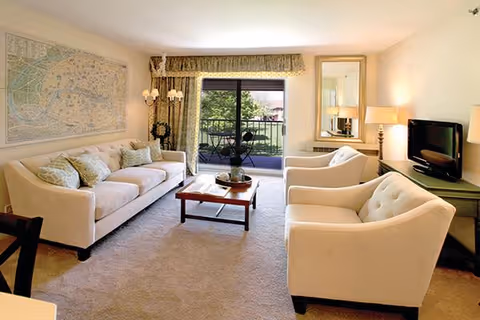 A bright living room with a beige carpet, a cream-colored sofa with patterned cushions, two matching armchairs, a wooden coffee table, a TV on a green console, a floor lamp, and a large window with sliding glass doors leading to a balcony with outdoor seating.