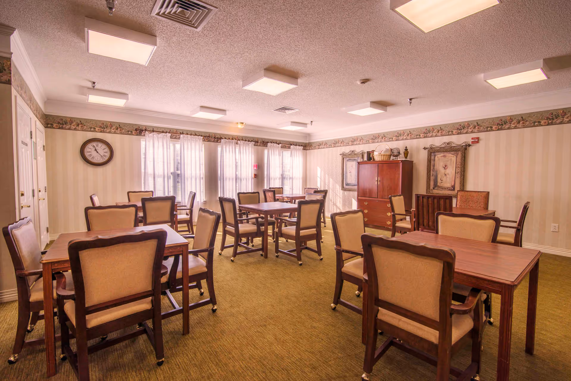 A well-lit dining room with several wooden tables and cushioned chairs arranged neatly. The room has beige walls with a floral border near the ceiling, a green carpeted floor, and large windows with sheer white curtains allowing natural light to enter. There is a clock on one wall and framed artwork along with a wooden cabinet on another wall.