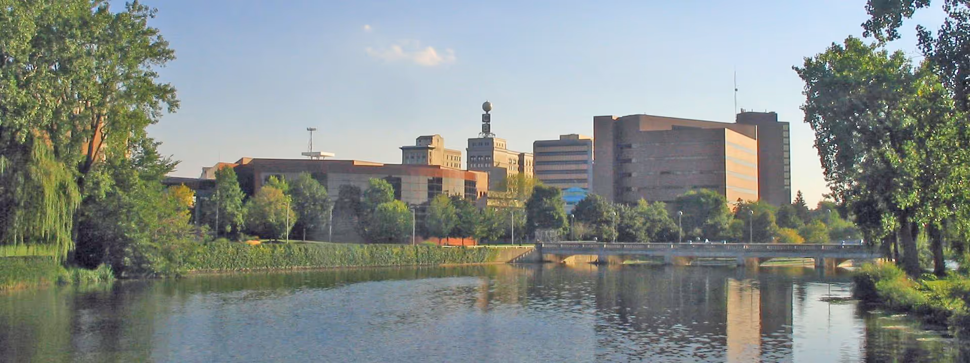 A calm lake with tree-lined banks and a bridge reflecting a row of mid-rise city buildings under a clear sky.