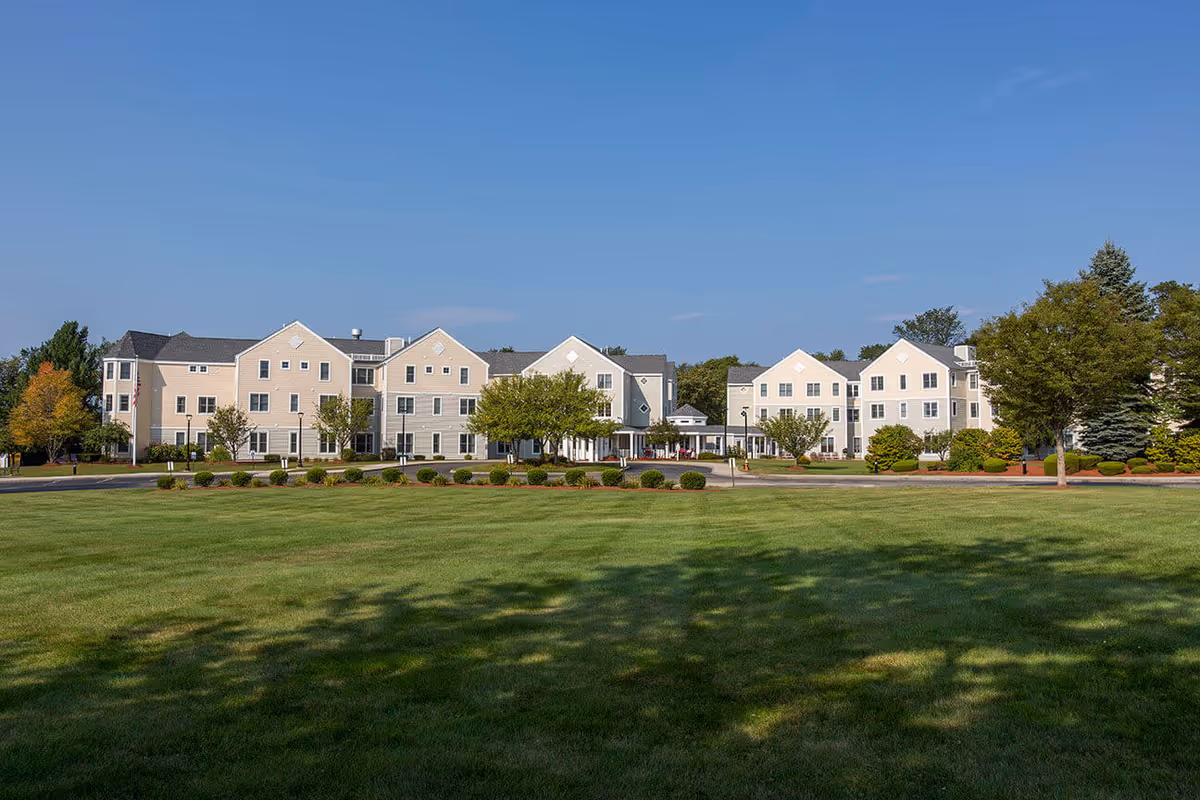 Exterior view of Benchmark at North Andover, a large beige multi-story senior living facility with multiple peaked roofs, surrounded by green lawns, trees, and shrubs under a clear blue sky.