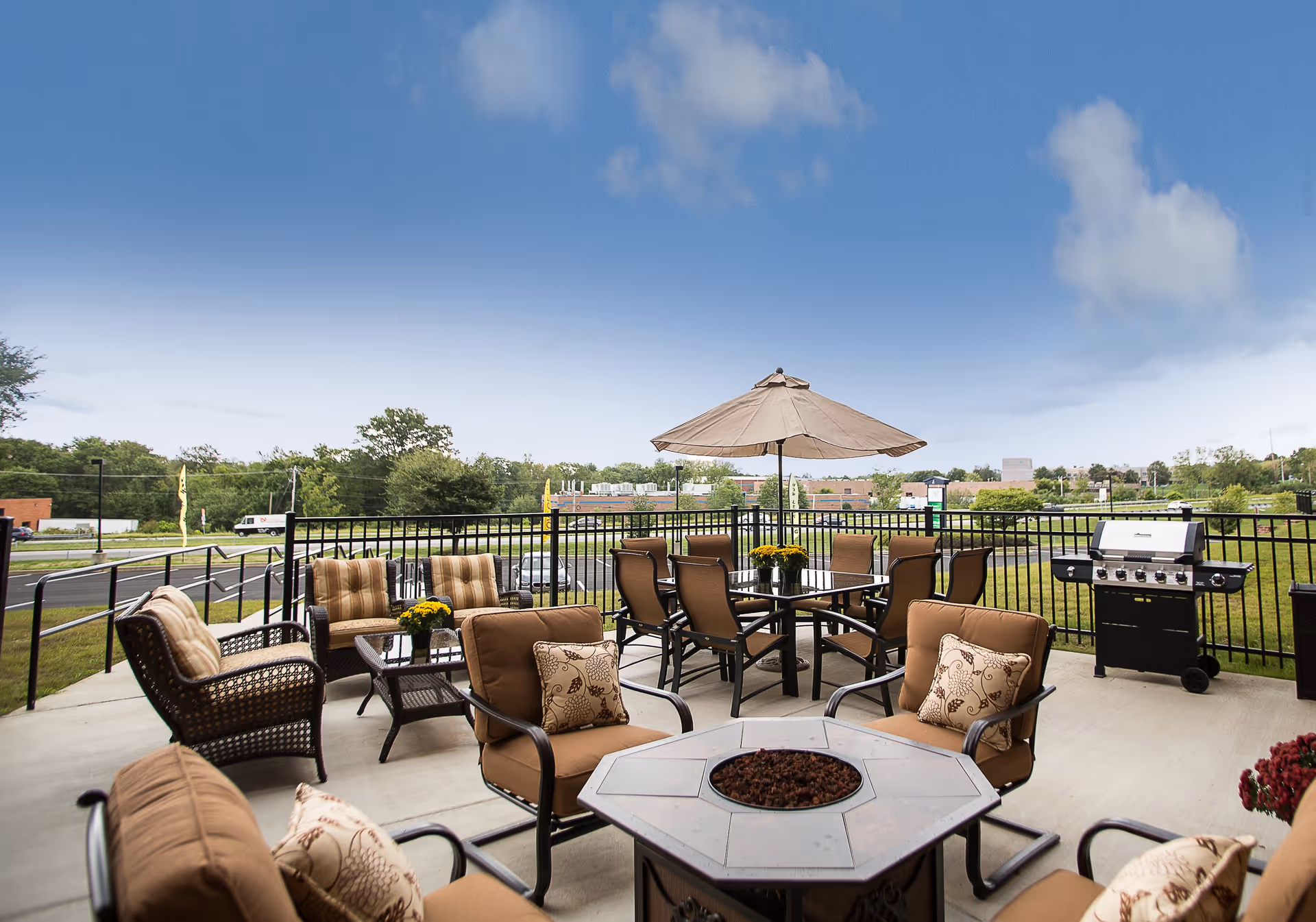 Outdoor patio with cushioned chairs, tables, an umbrella, a fire pit and a grill overlooking a grassy area and road.