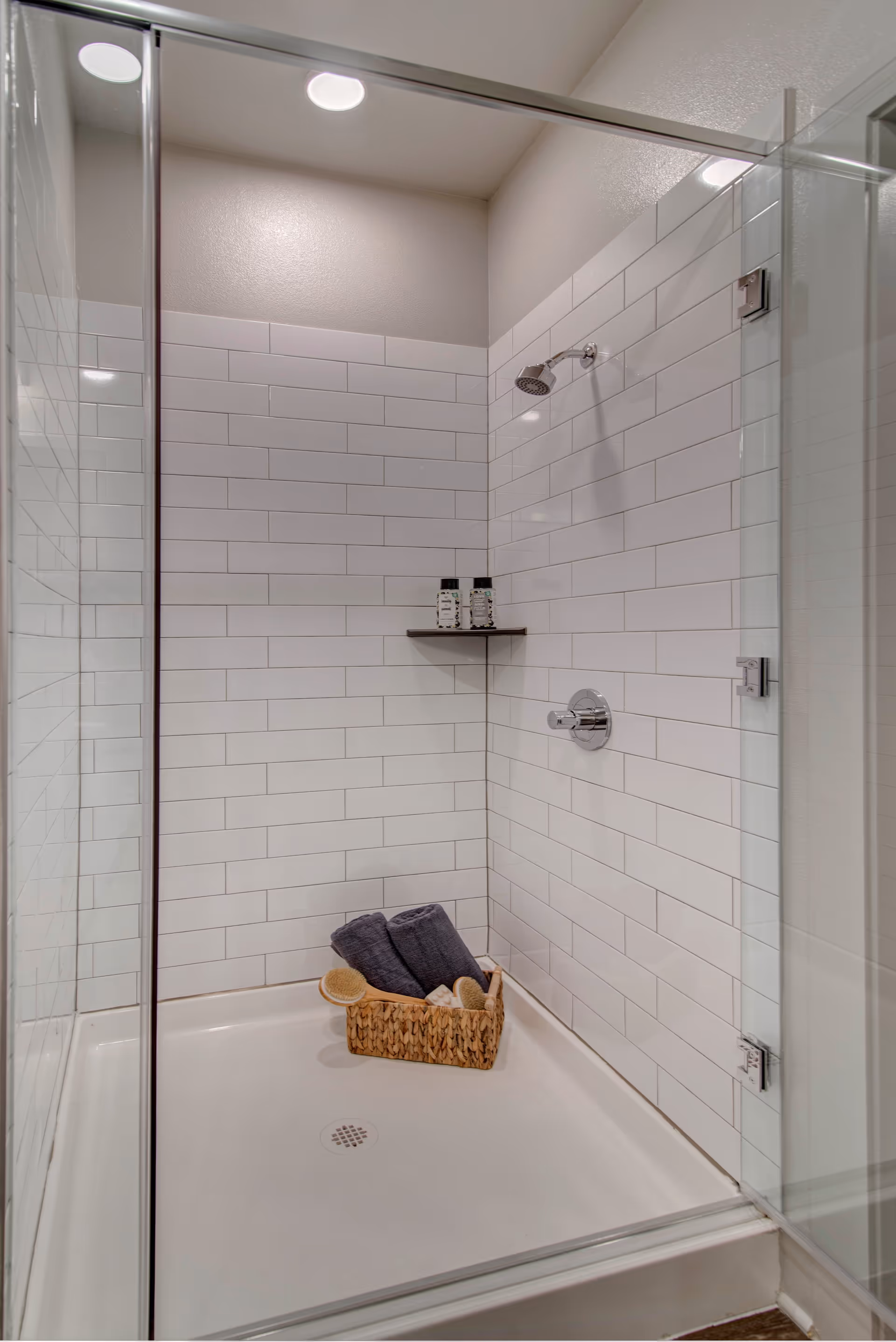 A clean, modern shower with white subway tile walls and a glass door. Inside the shower, there is a small corner shelf holding two bottles of shower products. On the shower floor, there is a woven basket containing two rolled dark gray towels and some bath brushes.