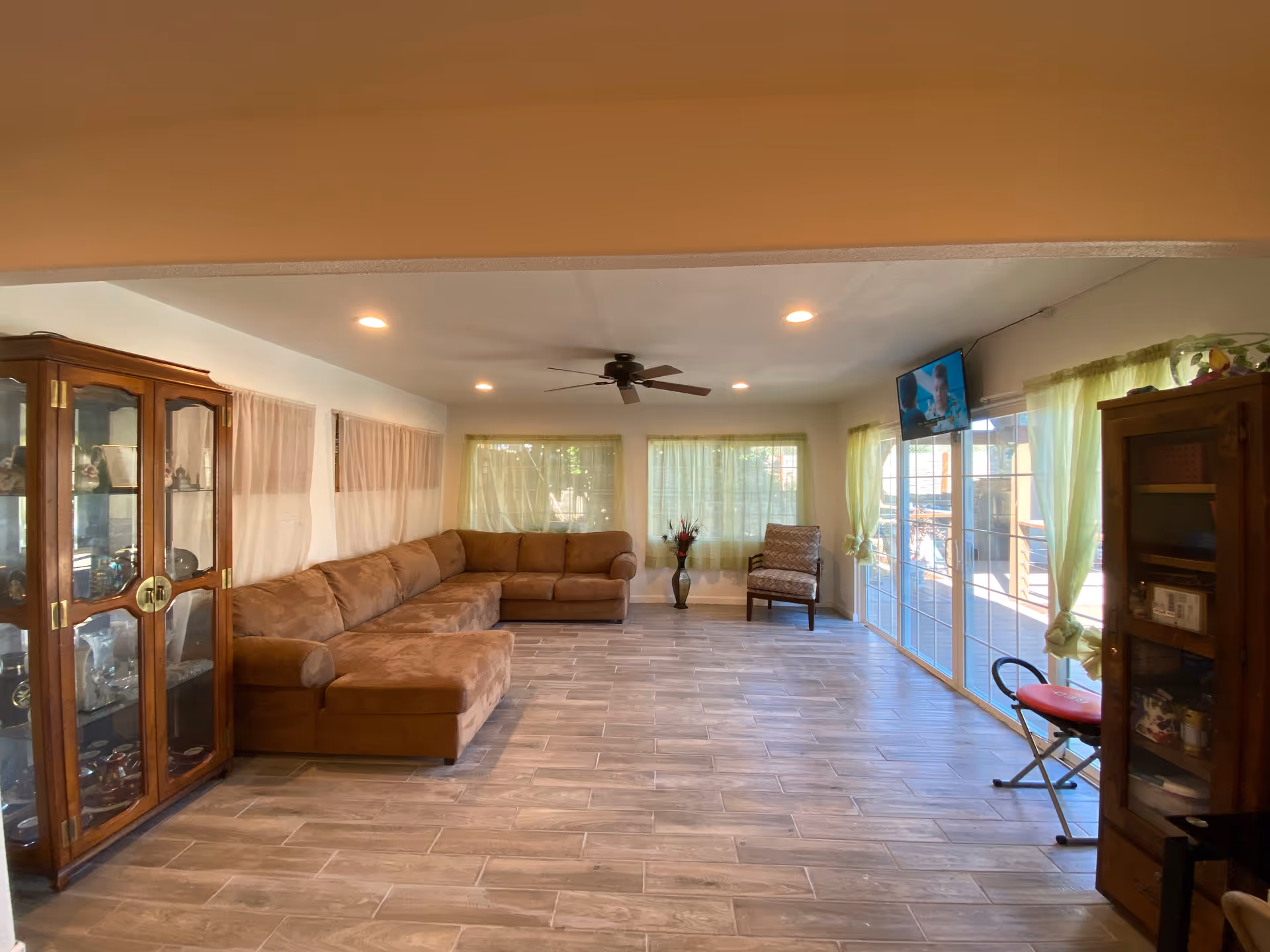A spacious living room with a large brown sectional sofa along the left wall, a single armchair near the windows, and a flat-screen TV mounted on the right wall. The room has large windows with light green curtains, a ceiling fan with lights, and wooden display cabinets on both sides. The floor is tiled with a wood-like pattern.