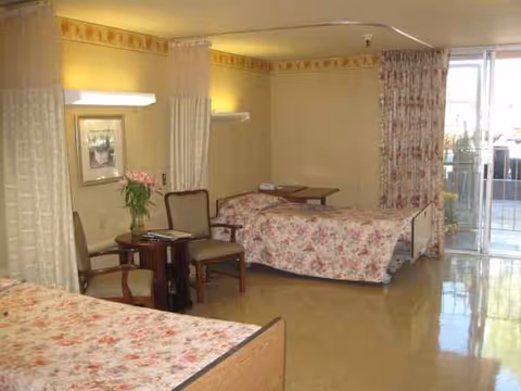Two beds with floral quilts, chairs, and a small table in a patient room.