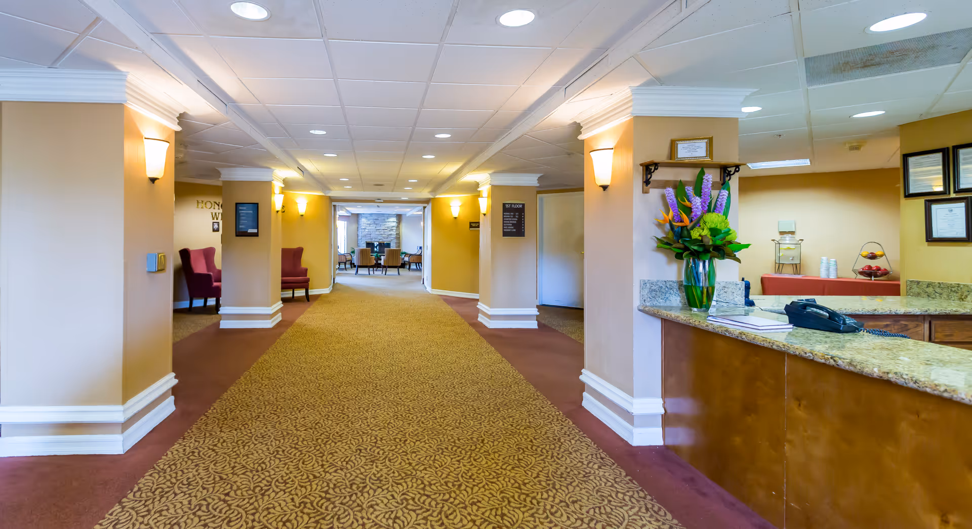 Interior hallway of a senior living facility with beige walls, patterned carpet, and ceiling lights. On the right side, there is a reception desk with a granite countertop, a telephone, a book, and a vase with colorful flowers. In the background, there are chairs and a stone fireplace in a common sitting area.
