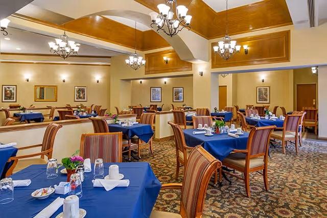 A spacious dining room with multiple tables covered in blue tablecloths, each set with white napkins, glasses, and small flower arrangements. The room features patterned carpet, wooden chairs with striped upholstery, chandeliers hanging from a high ceiling with wooden accents, and framed artwork on cream-colored walls.