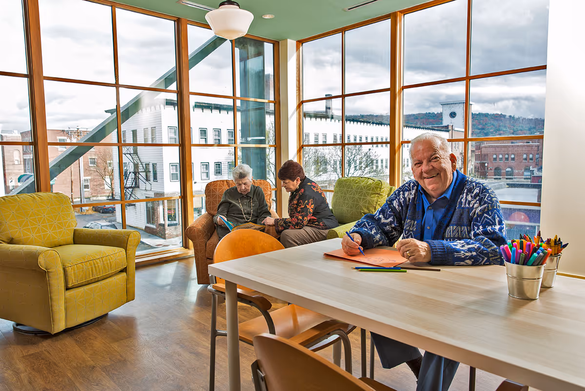 A bright room with large windows showing a cityscape outside. Inside, an elderly man is sitting at a table drawing with colored pencils and smiling at the camera. In the background, two women are seated in armchairs, one reading a book while the other looks on. The room has wooden floors and comfortable chairs in yellow, orange, and green patterns.