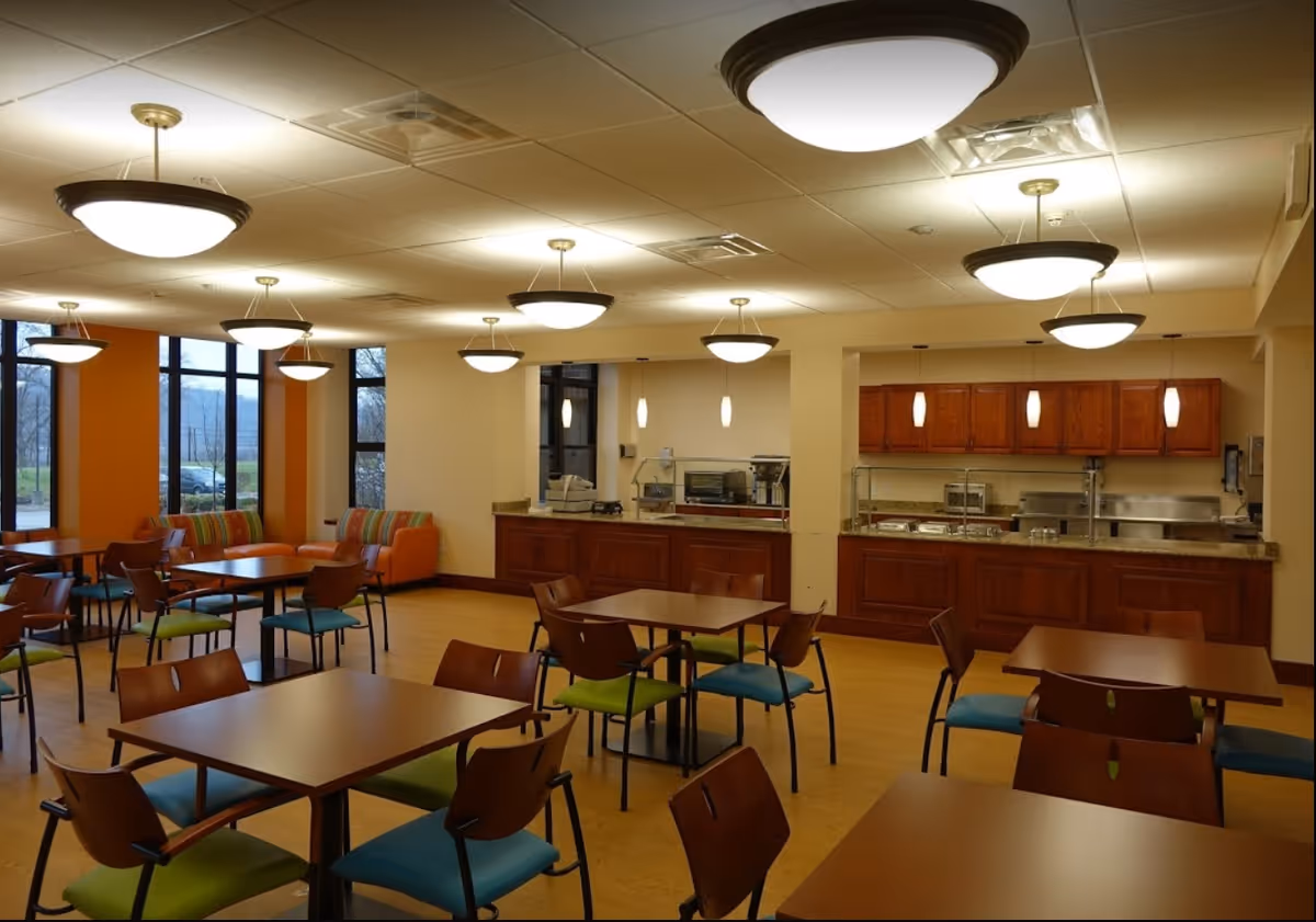 A dining area in a senior living facility with multiple tables and chairs, colorful cushioned seating along the windows, and a serving counter with kitchen appliances and cabinets in the background. The room is well-lit with ceiling lights and large windows letting in natural light.