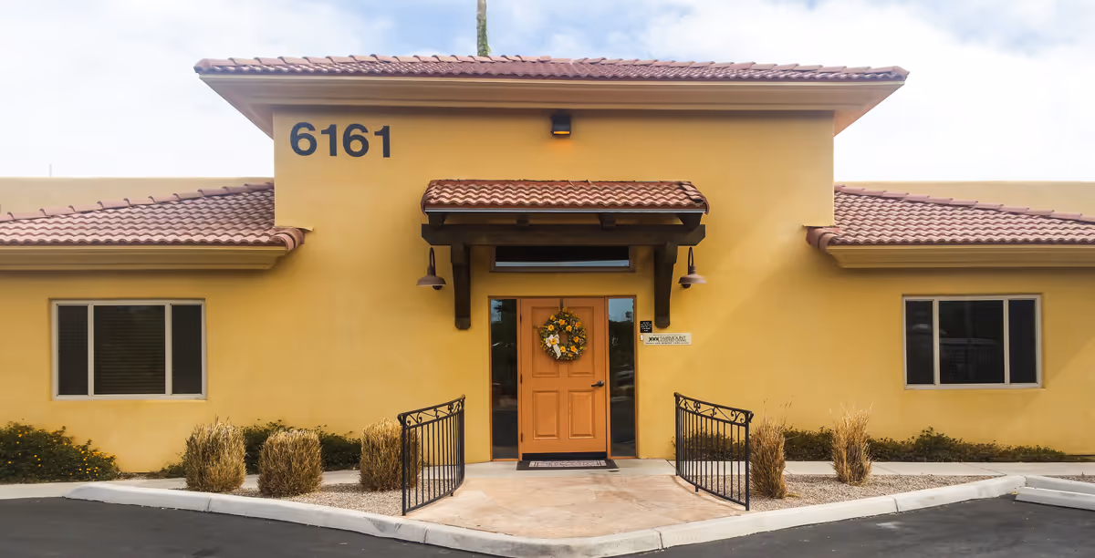 Front exterior view of a single-story building with a tan stucco finish and a red tile roof. The building has a central entrance with a wooden door decorated with a floral wreath, flanked by two windows. The address number 6161 is displayed prominently above the entrance. There are small bushes and landscaping in front of the building, and a paved driveway or parking area in the foreground.