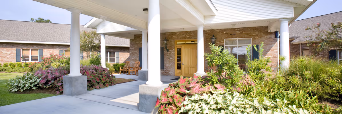 Entrance of a healthcare facility with a covered walkway supported by white columns, surrounded by well-maintained greenery and bushes, with a brick building and wooden door in the background.