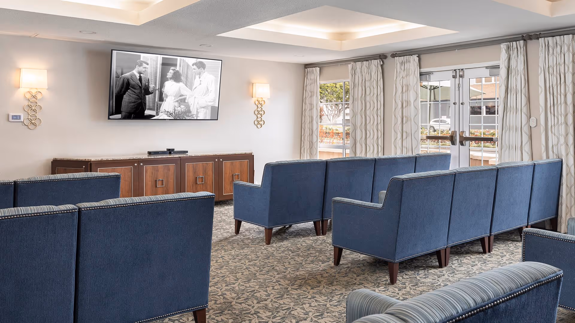 A cozy living room area with multiple blue upholstered chairs arranged in rows facing a wall-mounted flat-screen TV displaying a black and white movie scene. The room has patterned carpet flooring, wall sconces with warm lighting, large windows with patterned curtains, and a wooden cabinet beneath the TV.