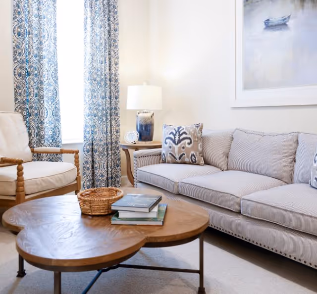 A cozy living room with a striped sofa adorned with patterned pillows, a wooden armchair with light cushions, a wooden coffee table with books and a small basket, blue patterned curtains, a side table with a blue and white lamp, and a framed painting of a boat on the wall.
