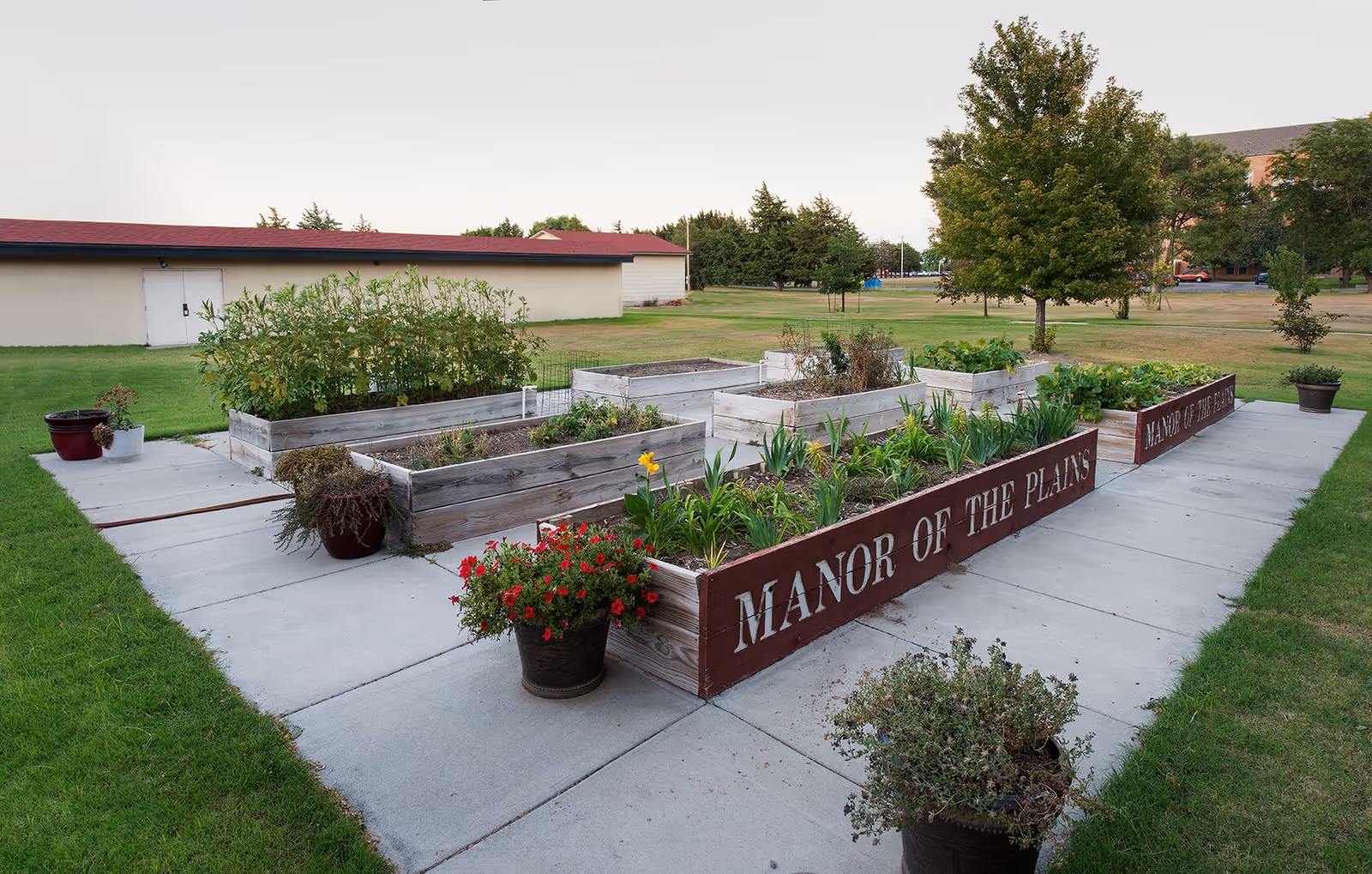 Raised wooden planter boxes and potted flowers on a concrete patio reading 'Manor of the Plains' with lawn and trees in the background.