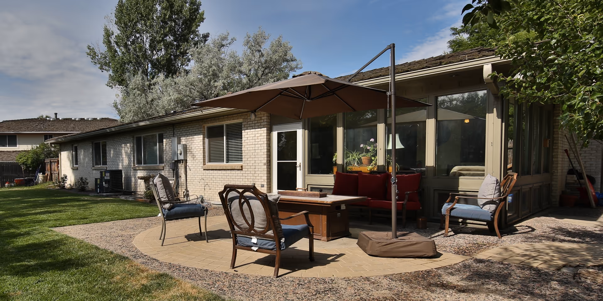 Outdoor patio area at Lighthouse Assisted Living Inc - Newland featuring a circular paved seating area with cushioned chairs, a large umbrella, a fire pit table, and a red cushioned bench. The patio is adjacent to a brick building with large windows and surrounded by green grass and trees.