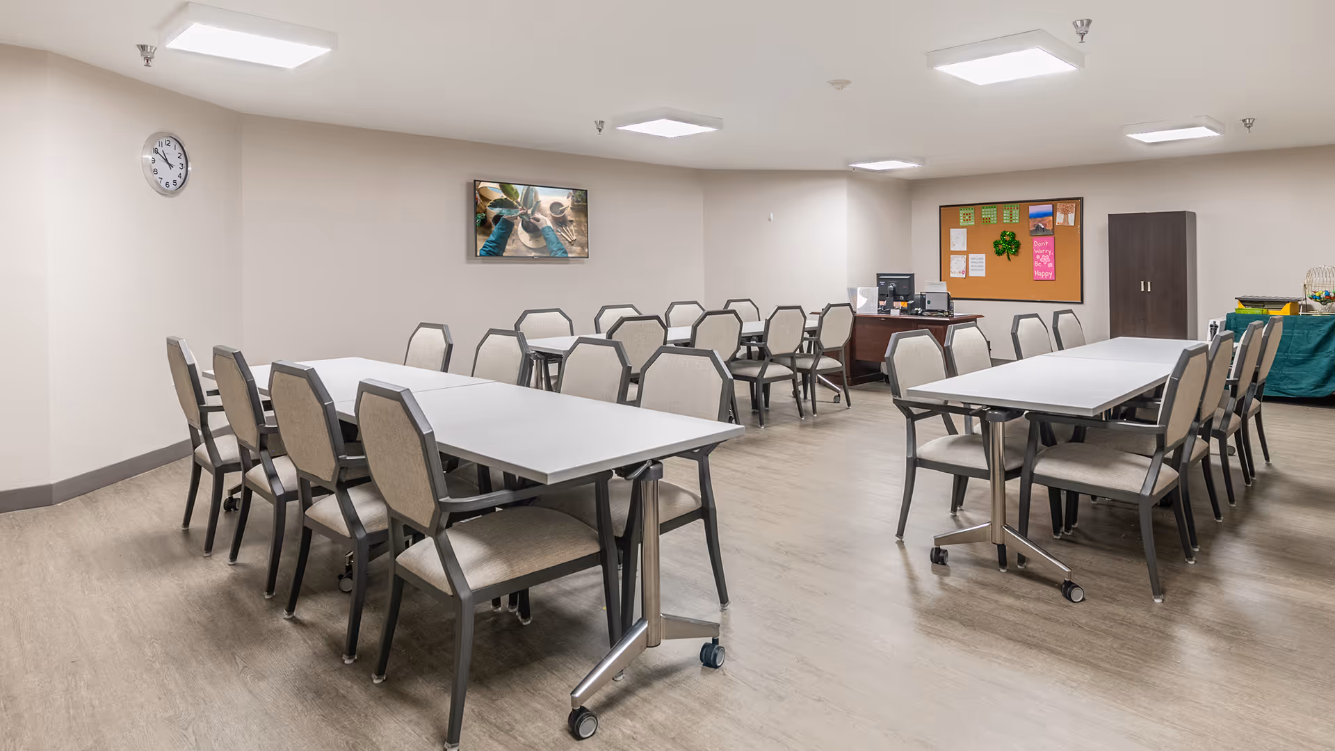 A clean and well-lit dining room with two long white tables surrounded by beige cushioned chairs. The room has light-colored walls and flooring, a clock on the wall, a bulletin board with various notices, a cabinet, and a table with refreshments in the corner.