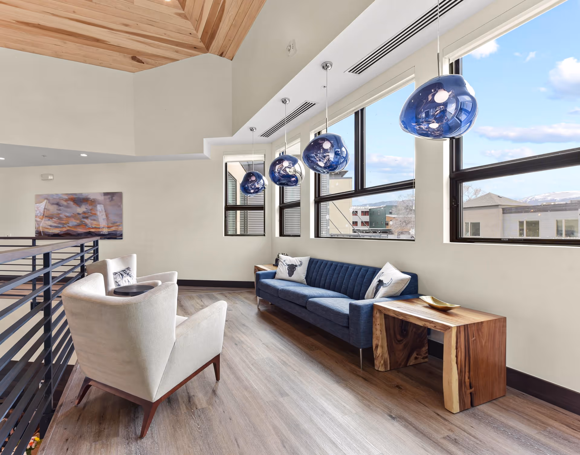 A modern senior living facility lounge area with a blue sofa, two white armchairs, wooden side tables, and large windows letting in natural light. Blue pendant lights hang from the ceiling, and a painting is visible on the far wall.