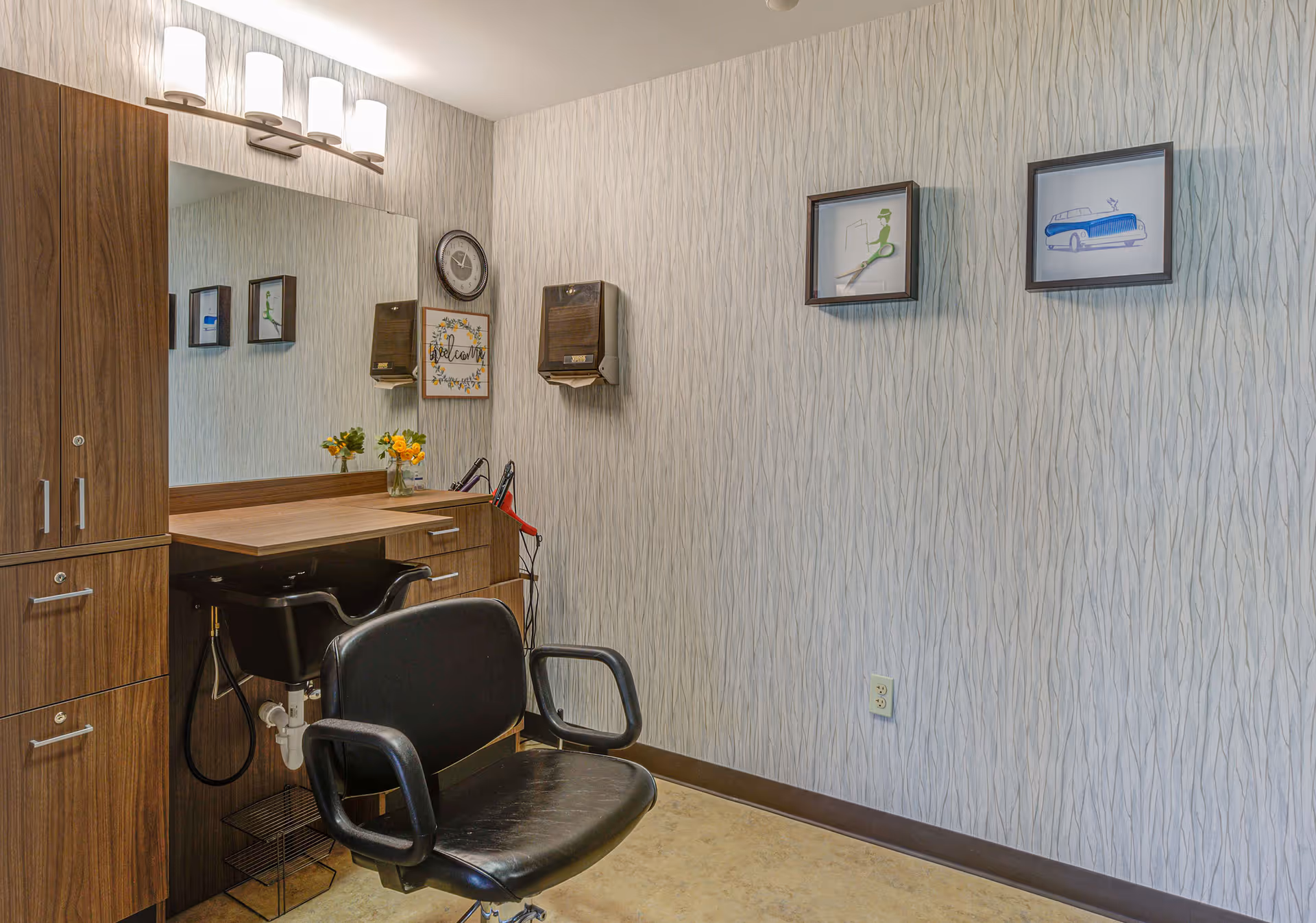 Interior view of a hair salon or grooming area with a black salon chair in front of a wooden counter and large mirror. The wall has light-colored textured wallpaper with two framed pictures and a clock. There are two wall-mounted dispensers and a small vase with yellow flowers on the counter.