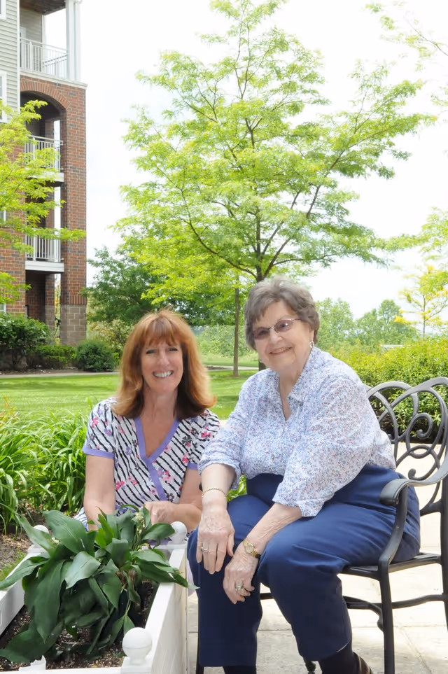 Two women sitting outdoors near a garden planter with green plants. One woman with red hair is kneeling beside the planter, smiling, and the other woman with gray hair and glasses is sitting on a metal chair, also smiling. Behind them is a green lawn, trees, and part of a brick building with balconies.