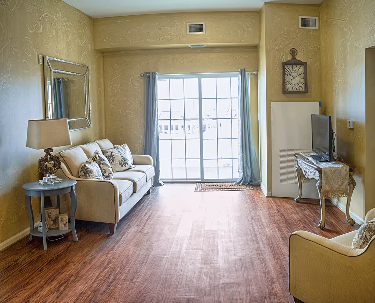 Bright living room with a sofa, armchair, side tables, decorative mirror and a TV by a sliding glass door.