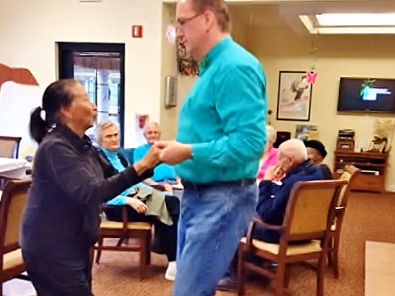 A man and a woman holding hands and dancing in a room with several elderly people seated in chairs watching them. The room has warm lighting and a television mounted on the wall.