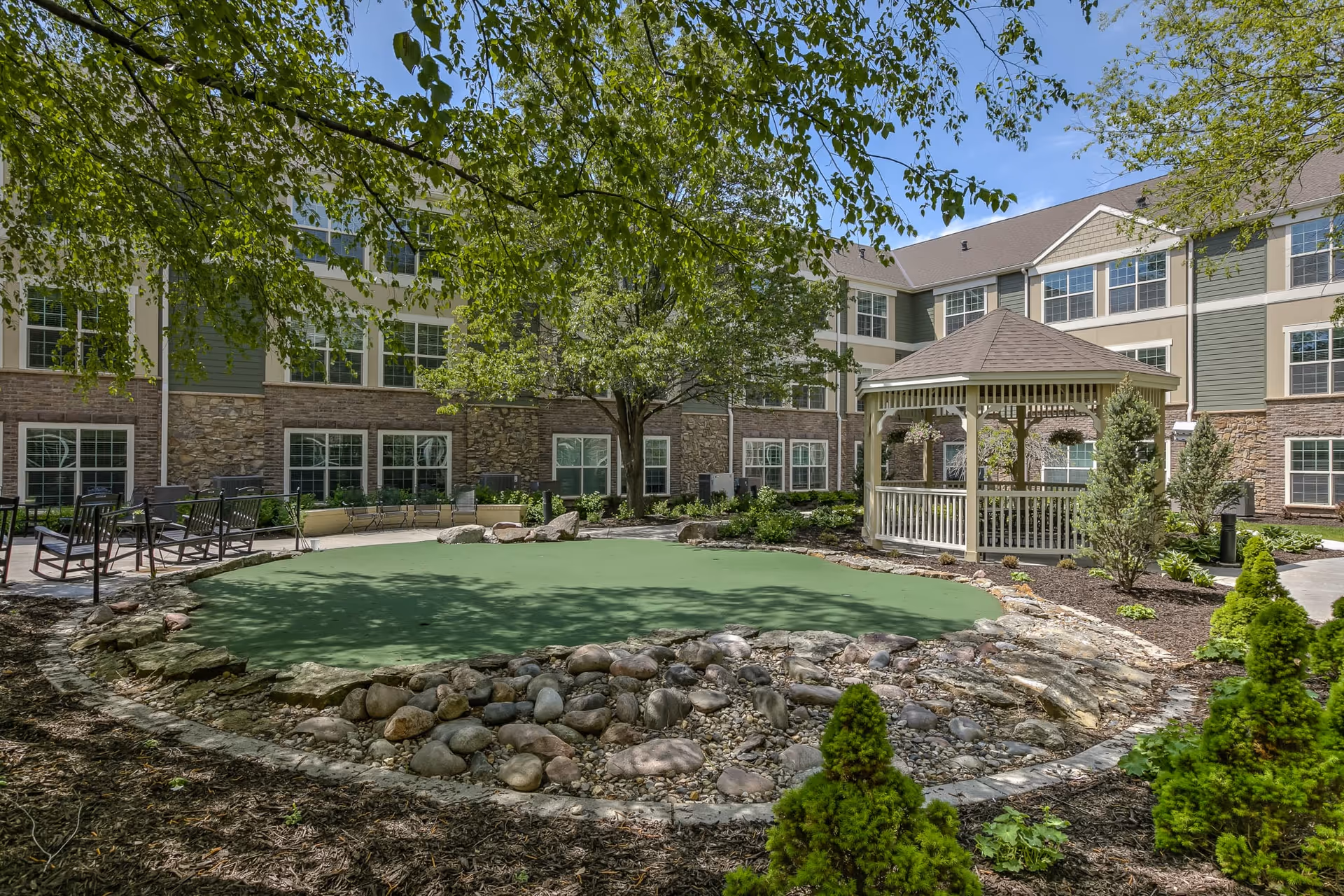 Outdoor courtyard area at Remington Heights Retirement Community featuring a green putting green surrounded by rocks and landscaping, a white gazebo, several trees, and a multi-story building with many windows in the background under a blue sky.