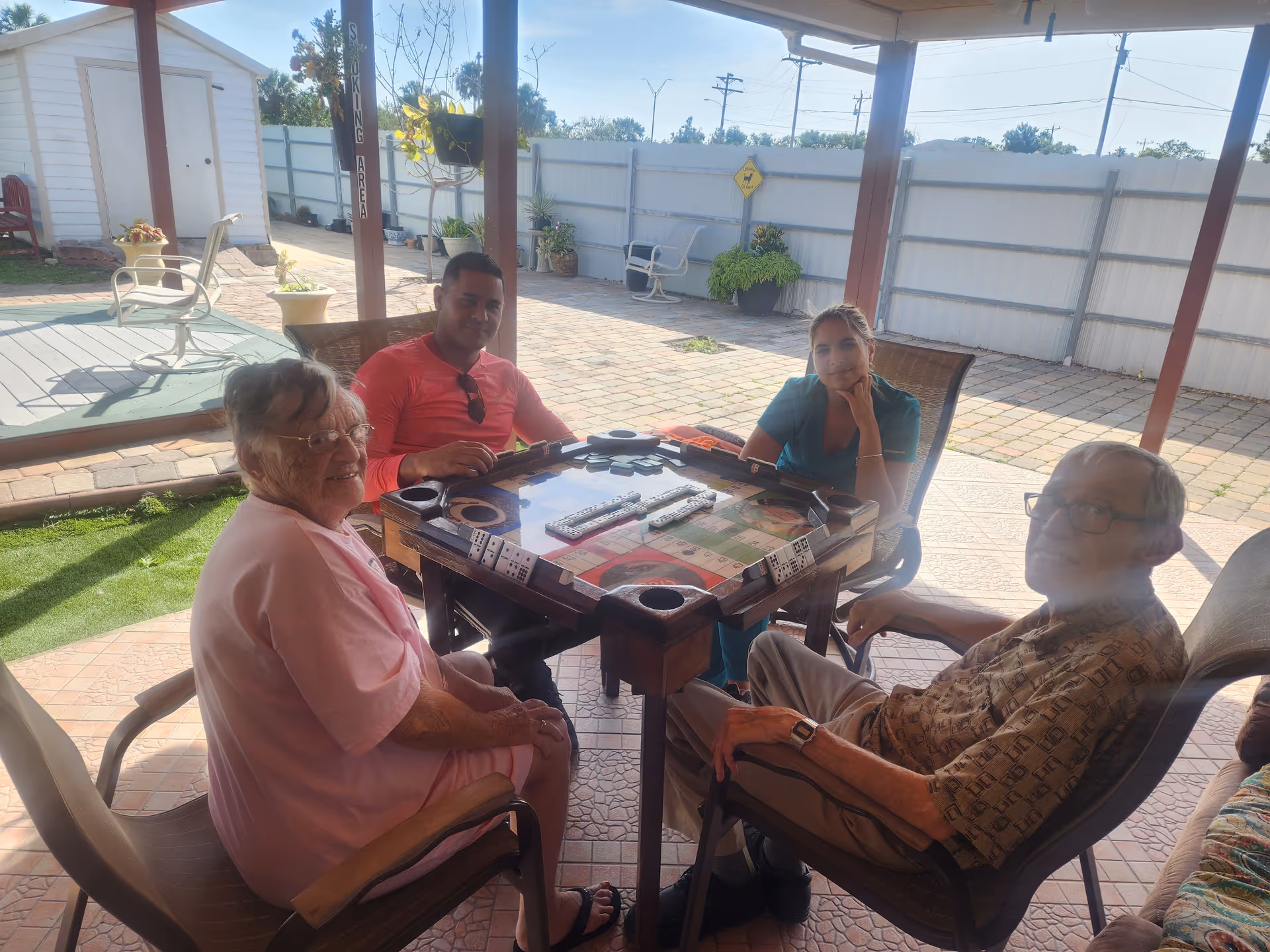Four people sitting around a square table playing a game of dominoes in a covered outdoor patio area. The group includes two elderly individuals and two younger adults. The background shows a fenced yard with potted plants and outdoor furniture.