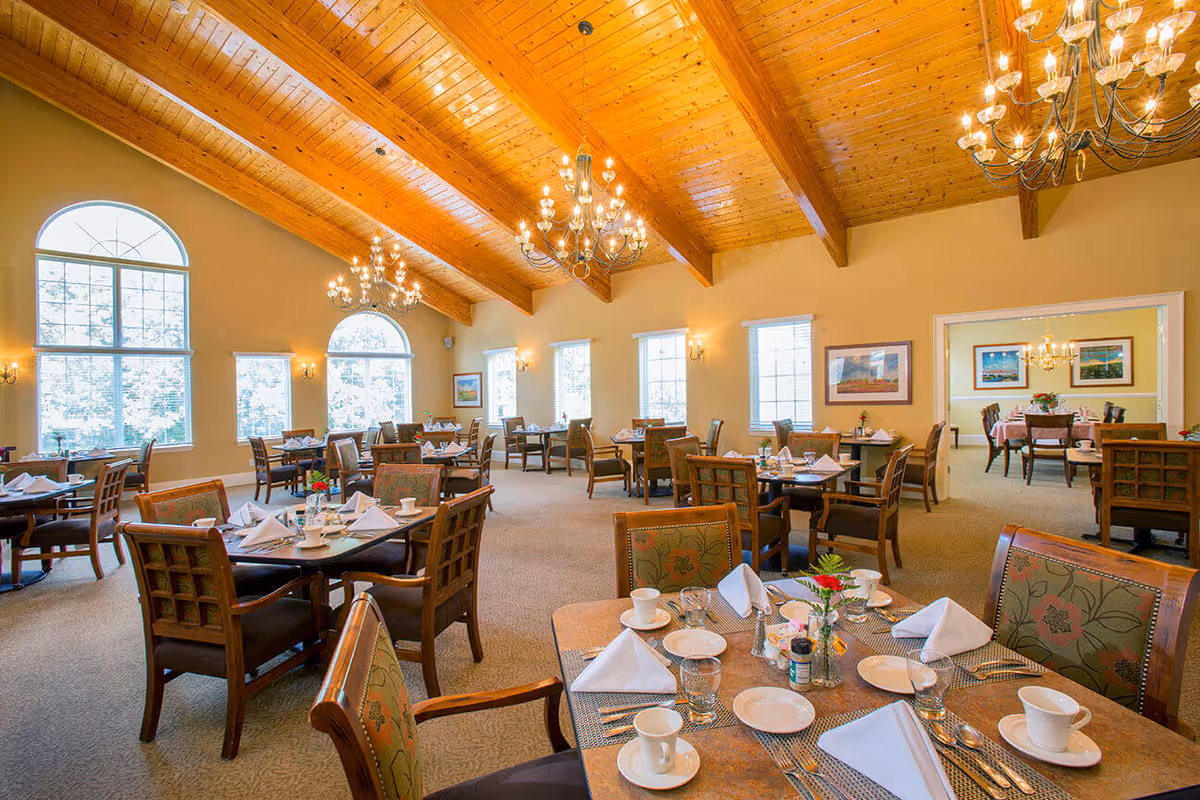 A spacious dining room with multiple tables set for a meal, featuring white napkins, cups, plates, and silverware. The room has large arched windows allowing natural light to fill the space, a high wooden ceiling with exposed beams, and elegant chandeliers hanging from the ceiling. The walls are painted a warm beige color and decorated with framed artwork. There is an adjoining room visible through a wide doorway, also set up with dining tables and chairs.