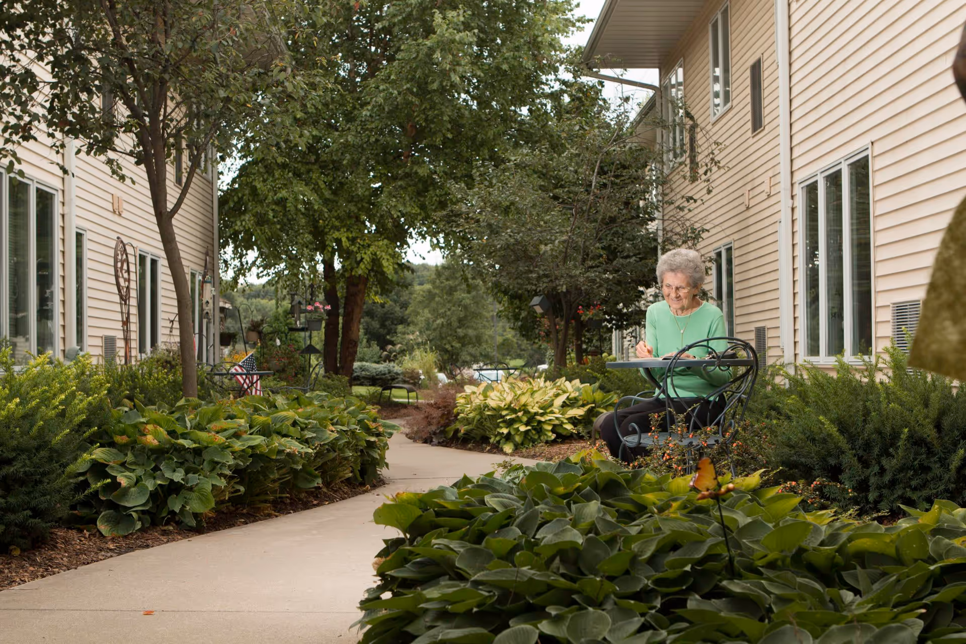 An elderly woman sitting at a small outdoor table in a garden area between two beige buildings. She is writing or drawing on a pad, surrounded by lush green plants and trees along a paved walkway.