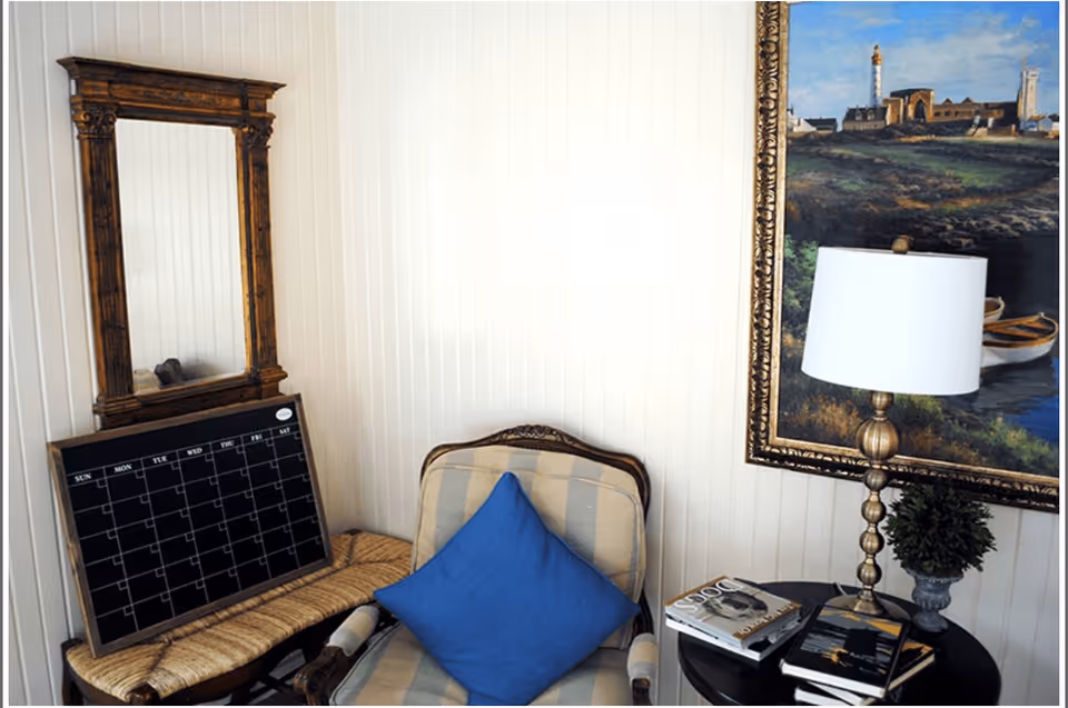 A cozy corner of a room featuring a cushioned armchair with a blue pillow, a small table with a lamp, books, and a small plant, a large framed painting on the wall, and a wooden-framed mirror above a woven bench with a blank weekly calendar board resting on it.
