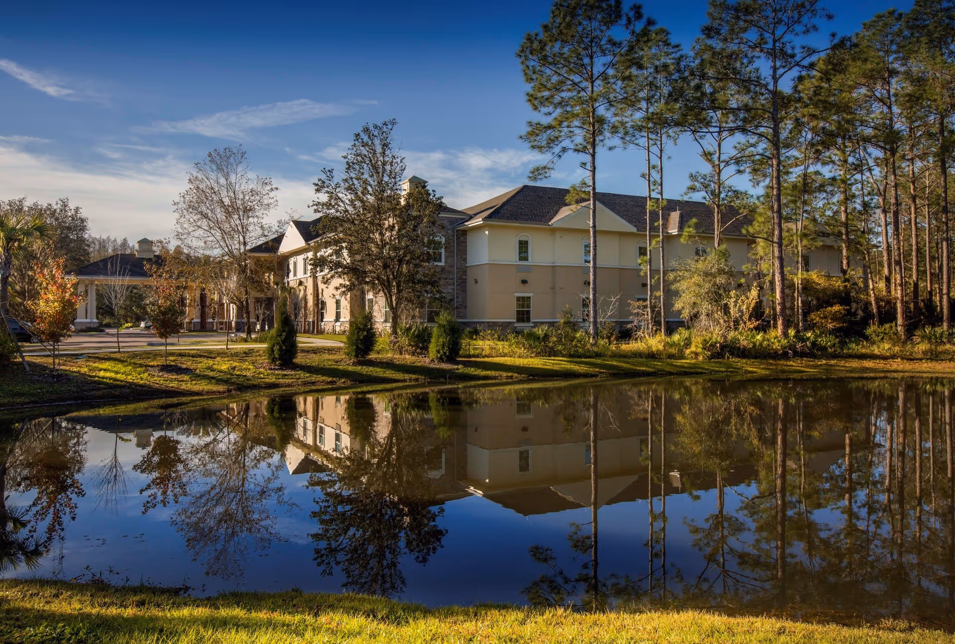 Exterior view of a senior living facility building named The Legacy at Highwoods Preserve, surrounded by trees and reflected in a calm pond under a blue sky.