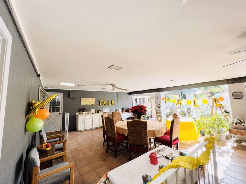 A bright and spacious indoor dining area decorated for a fall celebration with yellow and orange balloons and a banner. The room features a round table with a beige tablecloth surrounded by six upholstered chairs, a sideboard against a gray wall with the word 'FALL' spelled out in gold balloons, and several potted plants near large windows letting in natural light. Additional chairs and tables with decorations are visible, creating a welcoming atmosphere.