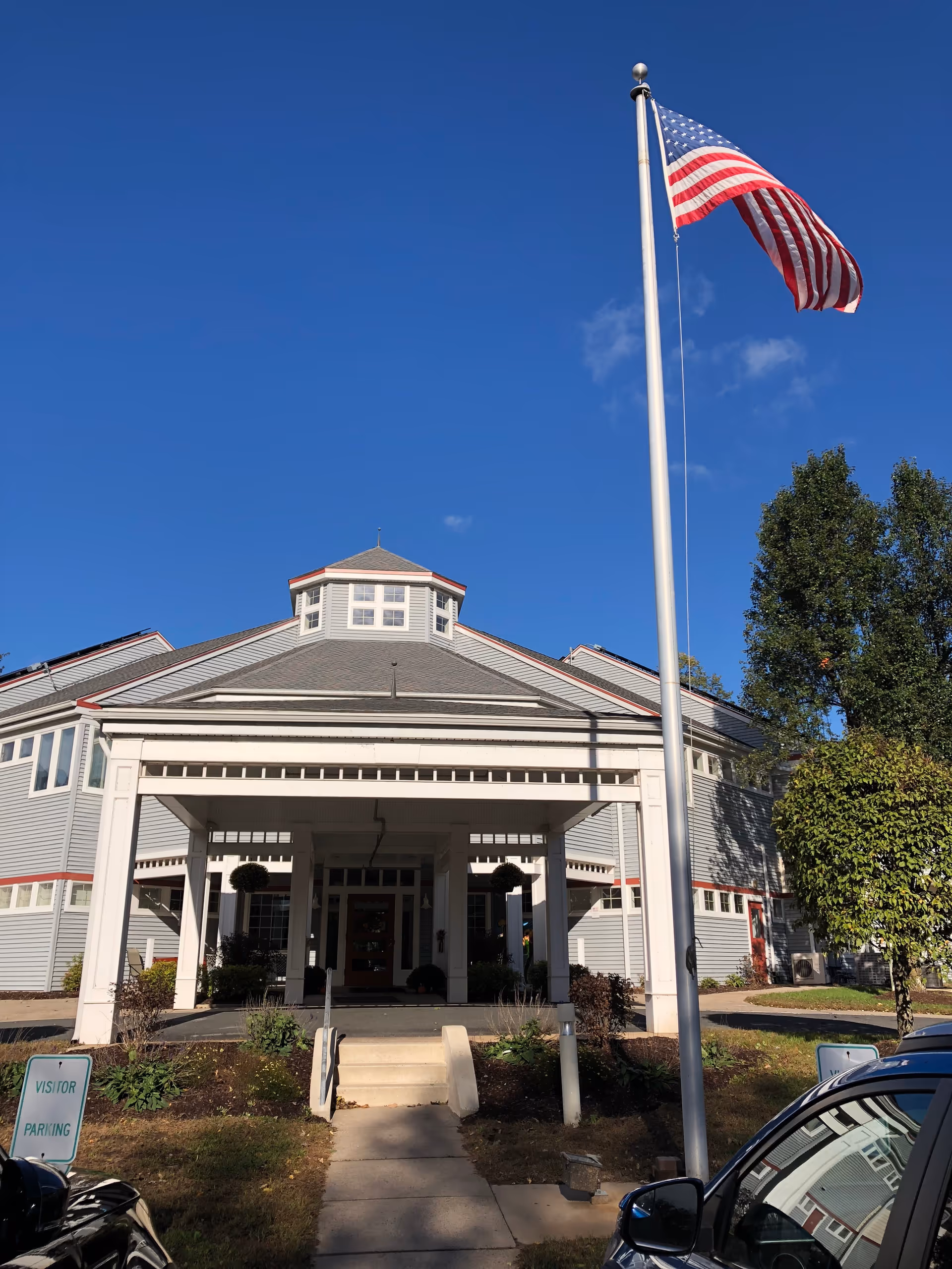 Front exterior view of a senior living facility building with a covered entrance, an American flag on a tall flagpole, and a clear blue sky. There are some trees and parked cars visible near the entrance.