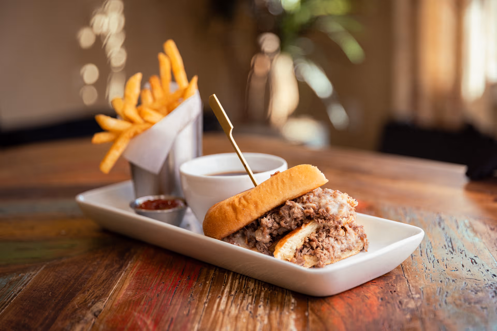 A close-up of a sandwich filled with chopped meat and melted cheese on a rectangular white plate, accompanied by a small metal container of French fries, a small cup of ketchup, and a white cup of dipping sauce, all placed on a rustic wooden table in a softly lit indoor setting.