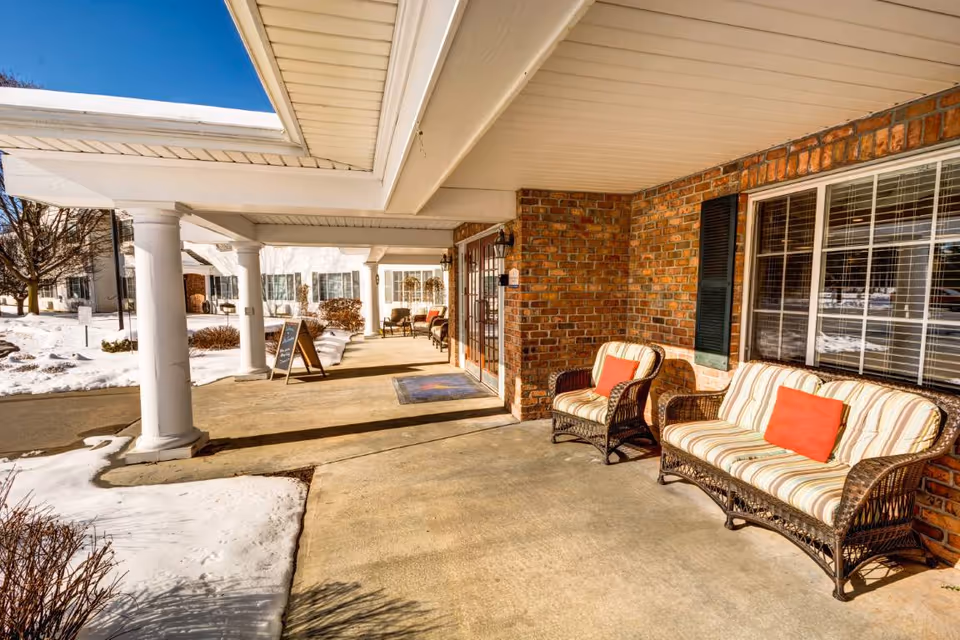 Covered outdoor patio area at Evergreen Place with wicker chairs and a loveseat featuring striped cushions and red pillows. The patio has white columns and a brick wall, with snow visible on the ground and a clear blue sky.