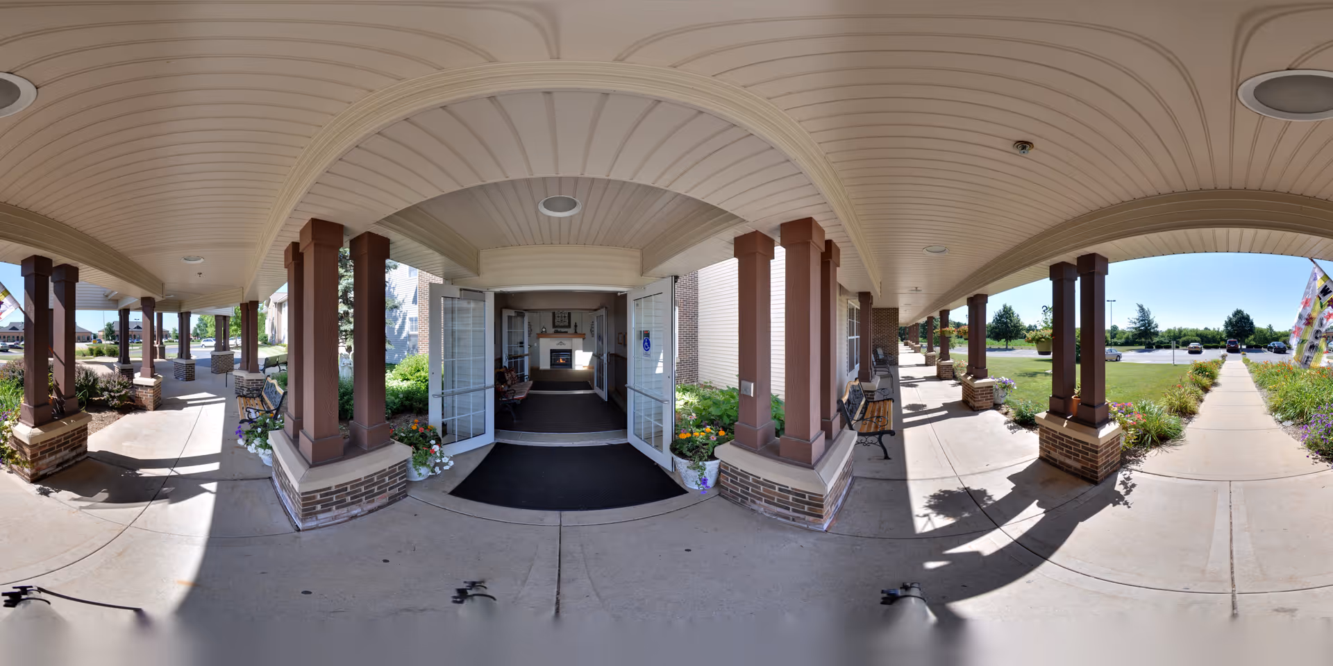Covered entrance walkway of a senior living facility with brick columns and a curved ceiling. The entrance has double glass doors leading inside, with benches and flower pots on either side. Outside, there is a sidewalk, landscaped greenery, and a parking area visible under a clear blue sky.