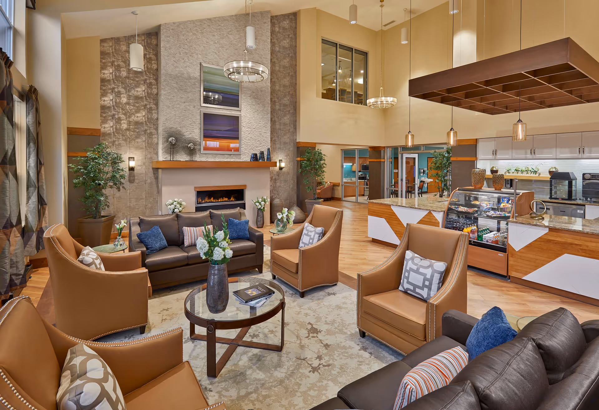 A spacious and well-lit living room area in a nursing home featuring a modern fireplace with decorative art above it. The room has a brown leather sofa and four tan leather armchairs arranged around a glass coffee table with a vase of flowers. The floor is wooden with a large area rug. In the background, there is a kitchen or snack bar area with a display case containing snacks and drinks, pendant lighting, and large windows overlooking other rooms.