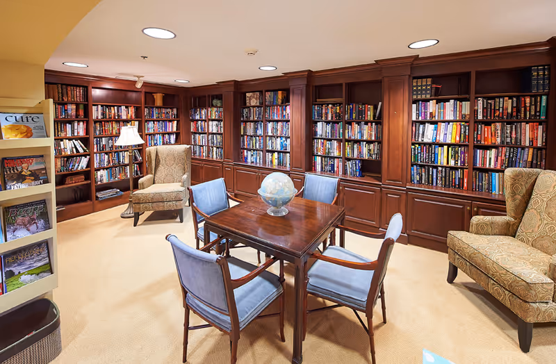 A cozy library room with wooden bookshelves filled with books along the walls, two patterned armchairs, a wooden table with four blue cushioned chairs around it, and a globe placed on the table. A magazine rack is visible on the left side of the image.