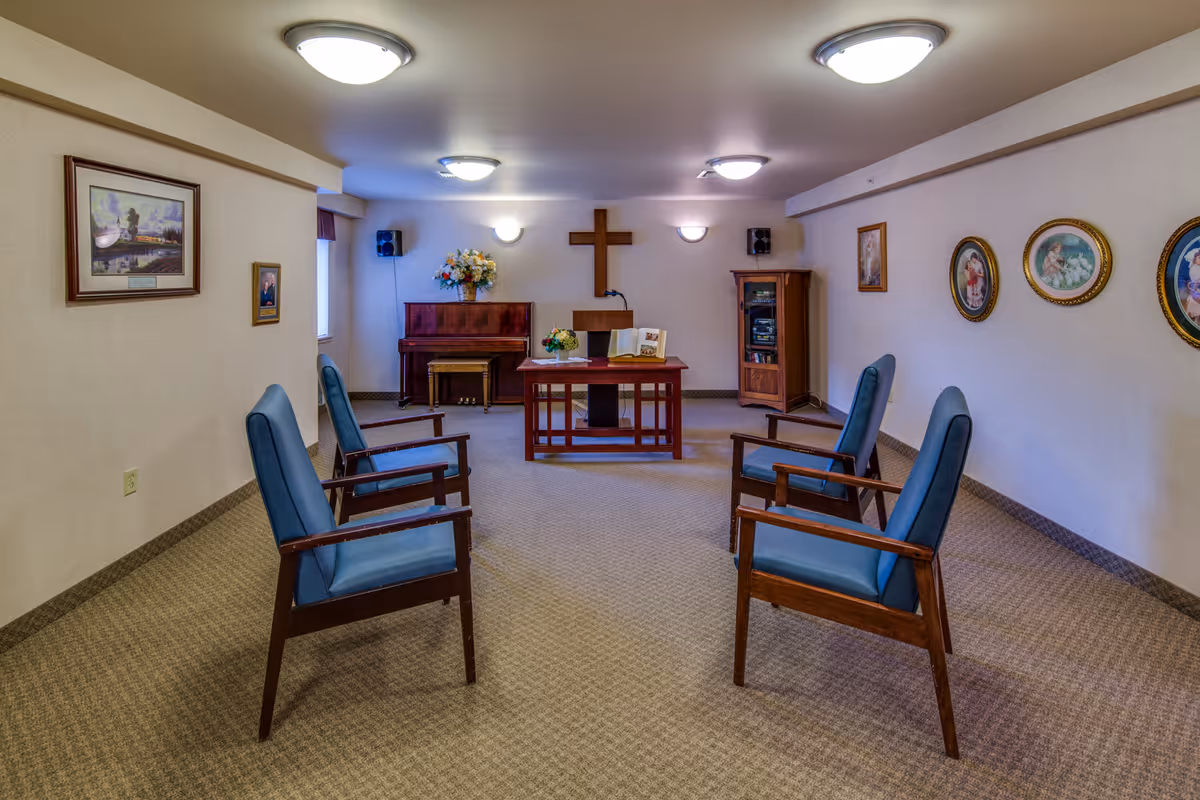 Small chapel-style meeting room with six blue upholstered chairs facing a wooden pulpit and cross, a piano, and framed wall art.