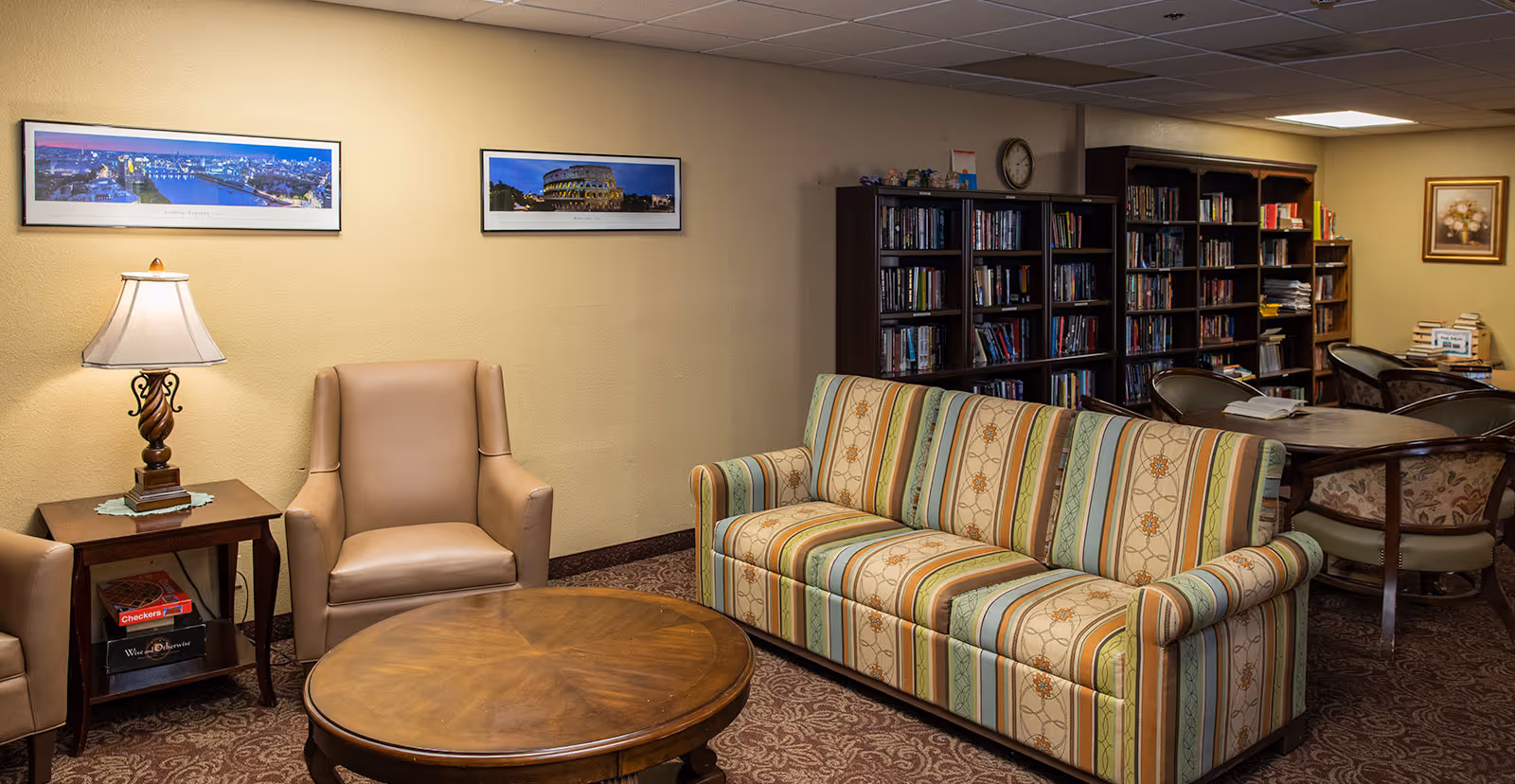 A cozy common area in a retirement community featuring a striped sofa, two beige armchairs, a round wooden coffee table, a side table with a lamp, and bookshelves filled with books. The walls have framed panoramic photos and a floral painting.