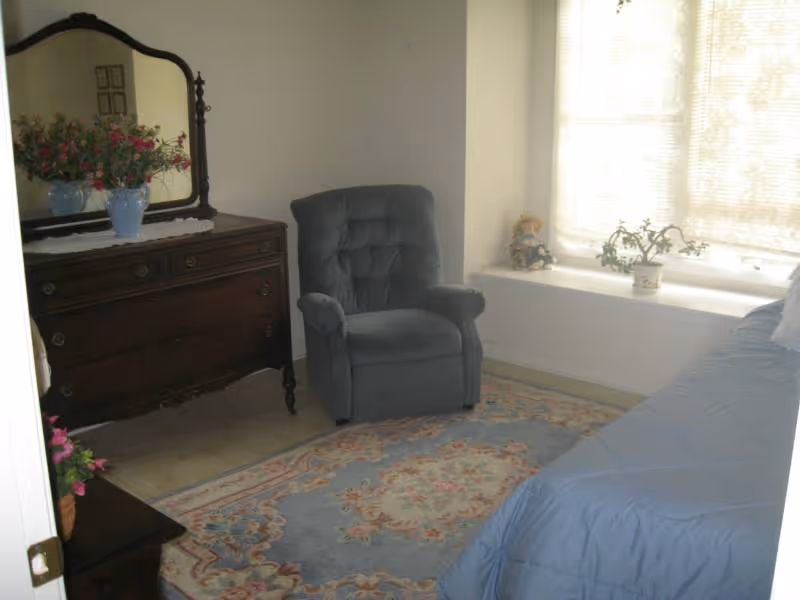 A cozy bedroom featuring a blue upholstered armchair, a wooden dresser with a large mirror and a vase of flowers on top, a floral area rug, a bed with a blue comforter, and a window seat with a potted plant and a doll.